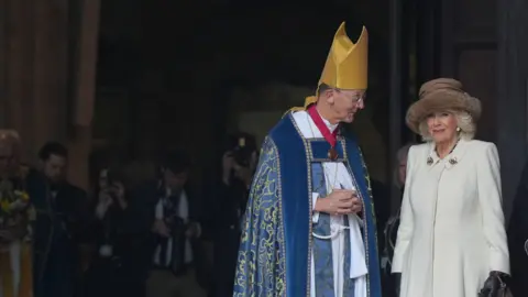 PA Bishop Inge, wearing a gold mitre and a blue robe, stands looking at The Queen who is wearing a cream-coloured coat and a brown hat.