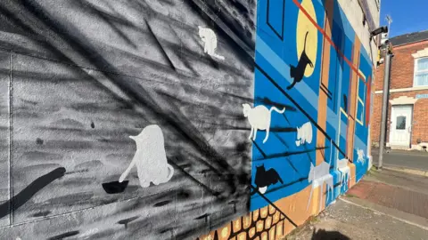 Murmur A mural shows cats who are sitting by food bowls. The background is black and blue.
