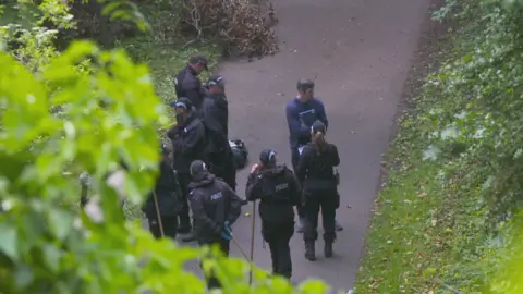 A group of police officers in black clothing with sticks standing on a concrete path with grass on either side. They are partially obscured by the leaves of a tree.