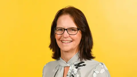 University of Suffolk Prof Paula Kersten is smiling at the camera. She is photographed against a yellow background and is wearing a silver/grey patterned top. She has shoulder-length brown hair and dark-rimmed spectacles.