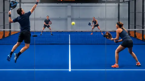 Two men and two women playing Padel wearing navy blue active wear and holding orange and blue padel rackets. They are playing in a padel court with a royal blue floor with orange and black nets. A sign above them reads Padel Chippenham.