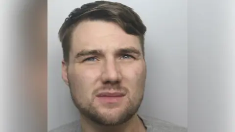 A mugshot of a man with short brown hair, stubble around his chin and a grey top.