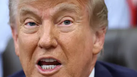 US President Donald Trump has his mouth open in a close up shot. His eyebrows are raised and he is wearing a blue suit jacket. 