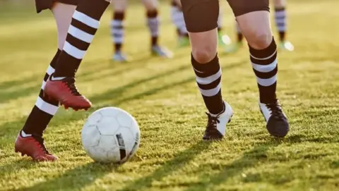 Getty Images Two sets of feat with socks and football boots on grass, between them they are kicking a white ball. It is a sunny day and there are blurry feet in the background