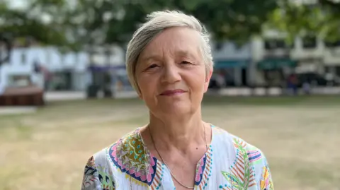 BBC Deputy Mary Le Hegarat, a woman with short grey hair. She is wearing a patterned blouse, which has a white base and multiple colours including pink, blue, green and yellow. She has a necklace around her neck. She is staring at the camera. Grass and trees are visible behind her and shops can be seen in the distance.