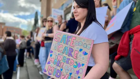Nikki Fox/BBC A student midwife stands with a cardboard poster decorated in pastel colours. It says in block writing "safe care needs more midwives". Other people are stood in the distance behind her. She has black, long hair and is looking thoughtfully to the distance. They are standing on the steps of city hall in Norwich.