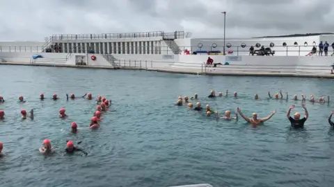 A number of swimmers with pink and yellow swim caps float with their heads above the water in a large open saltwater pool, with a white surround and some white-painted concrete changing booths on the far side from the camera.