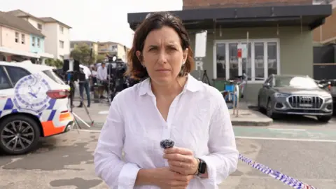 BBC's Katy Watson standing on road next to police tape and police car at Bondi Beach