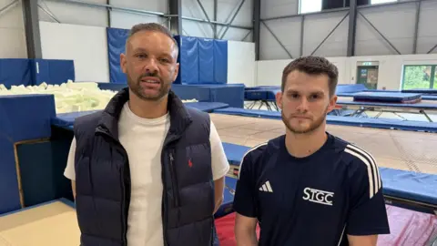 Two men are standing in a warehouse which contains a foam pit and a series of trampolines. One of the men is wearing a white t-shirt and navy blue gilet while the other is wearing a navy blue t-shirt with white lettering on it that reads STGC.