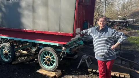 Rose Brash looks at the camera in a stripy jumper and red trousers, leaning against a traditional horse-drawn caravan