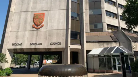 The side of a grey council building which has "Woking Borough Council" written on the side along with the council's emblem. 