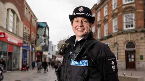 Essex Police Rachel Nolan smiles at the camera. It is a head-and-shoulders shot. She is wearing her uniform and police hat and is standing in a high street. The street is out of focus.