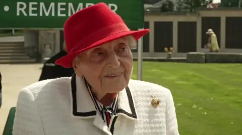 Suzanne sat on a chair at a D-Day remembrance event. She is wearing a smart cream jacket with black trim, a red hat and a gold broach. 