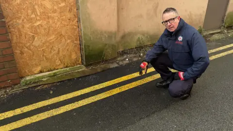 Firefighter Warren Topp in uniform crouching by double yellow lines where gang dug up road to join cables for cannabis farm
