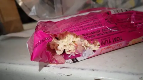 Liverpool City Council A pink packet of noodles on a shelf has a large hole at one end, with noodles visible.