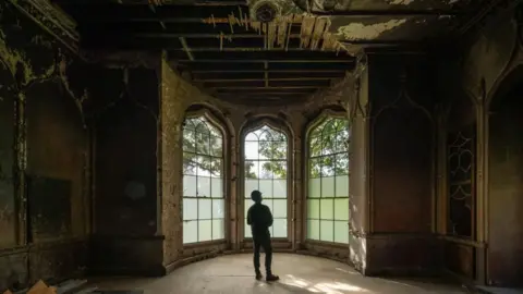 BHBT A man stood in an old, large room with dilapidated ceiling and a very large bay window