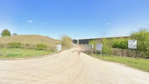 Cotesbach landfill site, near Shawell in Leicestershire. A road, flanked by an earth pile on the left and bushes to the right. A building is at the end of the road, which is bathed in sunlight.