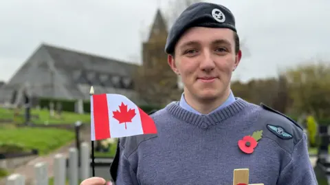 A young air cadet in blue uniform and regimental beret. He's wearing a poppy badge and holding a Canadian flag and standing in a cemetery. The church building is in the background. 