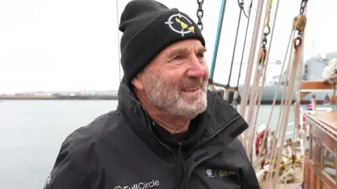  Dave Issitt looking to the right and smiling. He is wearing a black hat and black jacket. He is stood on the ship with the sea in the background.