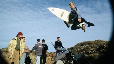 Carl Prechezer/FilmFour A fish eye camera view of five film cast members and a surfer who is jumping into the air with a surfboard under his arm.