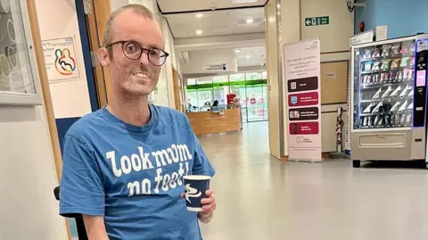Tom Staniford Tom Staniford is smiling as he sits in a wheelchair holding a paper cup. He is in a hospital corridor with a reception area behind him and a vending machine. He has short blond hair and is wearing glasses and a blue T-shirt with white writing that reads: Look mum no feet! 
