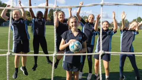 Sir Bernard Lovell Academy A close up of a group of girls aged about 12 on a football pitch dressed in navy blue kit. They are seen through the net and one in front is holding the ball, whilst the girls behind her are all jumping in the air with their fist held high.