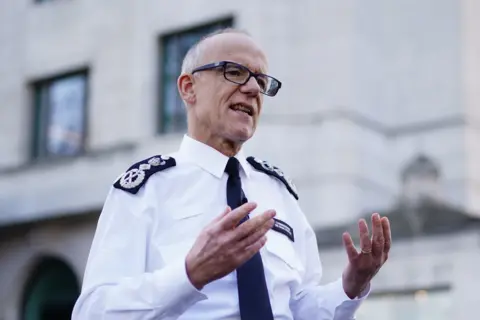Aaron Chown/PA Wire Sir Mark appears to be standing outside New Scotland Yard, addressing the media, he is wearing his police uniform and glasses and gesticulating with his hands.