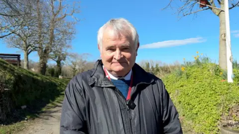 Connétable of Grouville, Mark Labey wearing a jacket and stood in a lane. He has short grey hair.