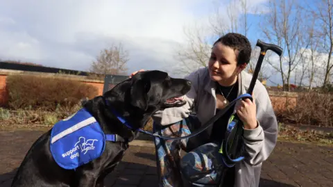 Support Dogs Elie-Mai Riddick crouches down to pet her black labrador dog.