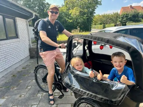 Henry Vanderpump Henry Vanderpump sits on an electric bike with a cart on the front carrying his two children