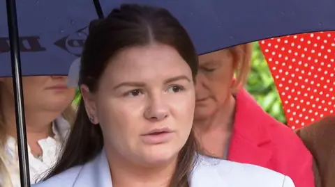 A young woman with long dark hair speaks to reporters, with several women standing behind her