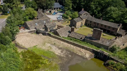 Aerial view of a historic stone-built mill complex beside a partially drained waterway. Several connected buildings with slate roofs are enclosed by stone walls, with a grassy courtyard at the centre. A parking area with multiple cars is visible behind the buildings, surrounded by mature trees and woodland. In the foreground, a muddy reservoir or mill pond edge and stone embankment can be seen, highlighting the site's industrial heritage setting.