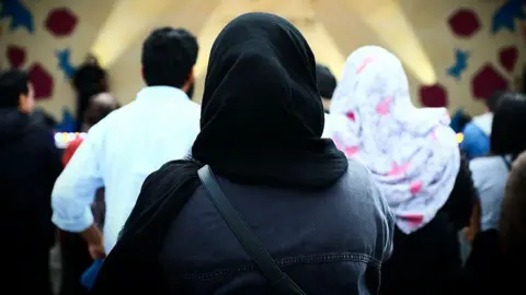 The backs of people, some of whom are wearing hijabs and jackets. They are facing the opposite direction to the camera and looking towards shafts of light. 