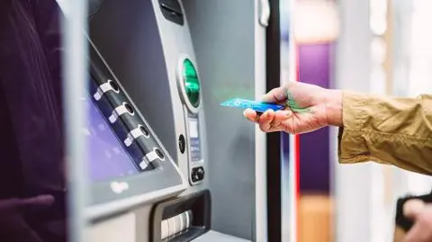 Getty Images A person reaches up to a ATM machine with their blue bank card in their hand.