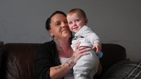 Kayleigh, a woman with dark hair and hoop earrings, sits on a sofa holding her baby. Kayleigh is looking into the camera and they are both smiling.