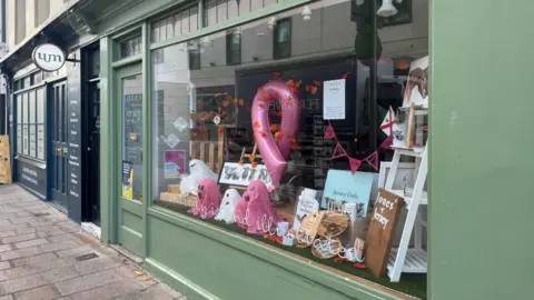A window display of a shop with pink and white ghosts and the pink cancer emblem as a balloon in the window.