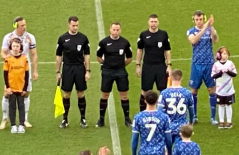 Carlisle United Players and children wearing ear defenders as teams come onto a football pitch