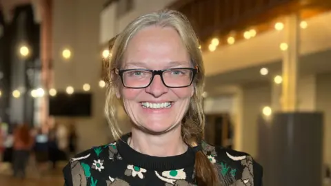 Carmel stands inside a large hall with high ceilings, warm decorative lighting, and a pointed architectural feature at the far end. 