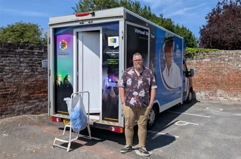 Adam Ross stands in front of a bus labelled "The Autism Reality Experience".  It is parked in a car park near a brick wall.