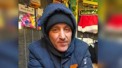 Chakib behind the counter of his shop he is wearing a dark blue coat and a dark woolen hat 