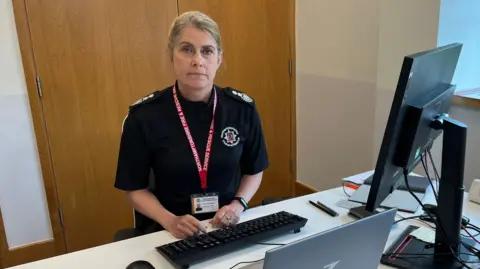 Steve Hubbard/BBC Northamptonshire Fire and Rescue Service chief fire officer Nikki Watson sitting behind a desk. She is wearing a black blouse and has an identity card on a red lanyard around her neck. There is a laptop, monitor and keyboard on the desk.