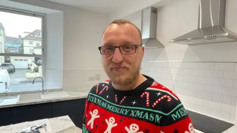 Andy Fletcher is pictured smiling at the camera and wearing a colourful Christmas jumper. He is wearing black glasses and is standing in one of the new kitchens. The kitchen has dark grey/blue units with white marble-effect surfaces and white tiles.