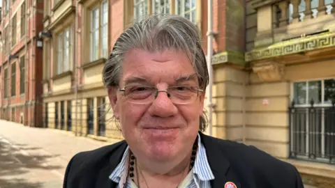 An image of Ged Mirfin outside County Hall in Preston. He is wearing a blue and white striped shirt and black blazer with a Fire Authority pin badge.  He is wearing glasses and smiling.