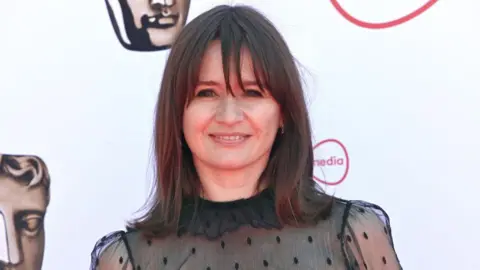 Emily Mortimer posing for photos at a Bafta awards ceremony. She is wearing a black chiffon top and has long dark hair. 