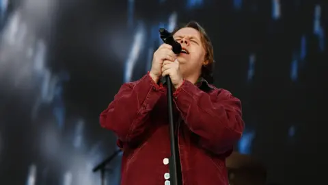 Lewis Capaldi performing on Radio 1's Big Weekend 2023. He sings into the microphone and is wearing a red shirt.