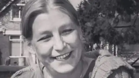 Family handout A black and white picture of Dawn Sturgess smiling at the camera, wearing a t-shirt, cardigan and a necklace. She is sitting on the grass in front of a house and a tree.