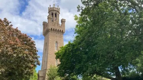 BBC Guernsey's Victoria Tower is seen through the trees while the sky in the background is blue with a lot of cloud coverage
