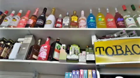 Two white shelves full of bottles of alcohol and vapes, next to a yellow sign advertising tobacco.