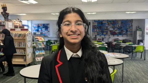 BBC/Kit Taylor A girl with black hair and clear glasses wearing school uniform and smiling. 