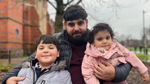 A man with dark hair and a beard is pictured holding two young children. On the left is a boy with dark hair wearing a grey coloured zip coat and on the right is a young girl with dark hair wearing a pink coat. In the background is a red brick building.